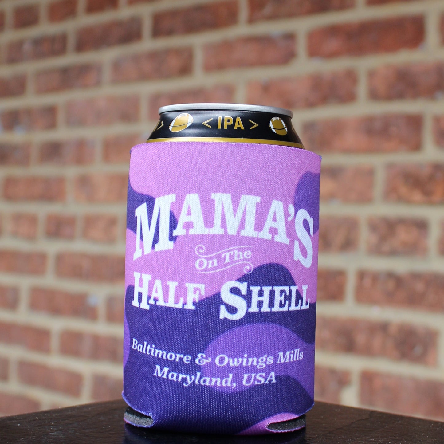 Purple koozie with Mama's logo against a brick wall.